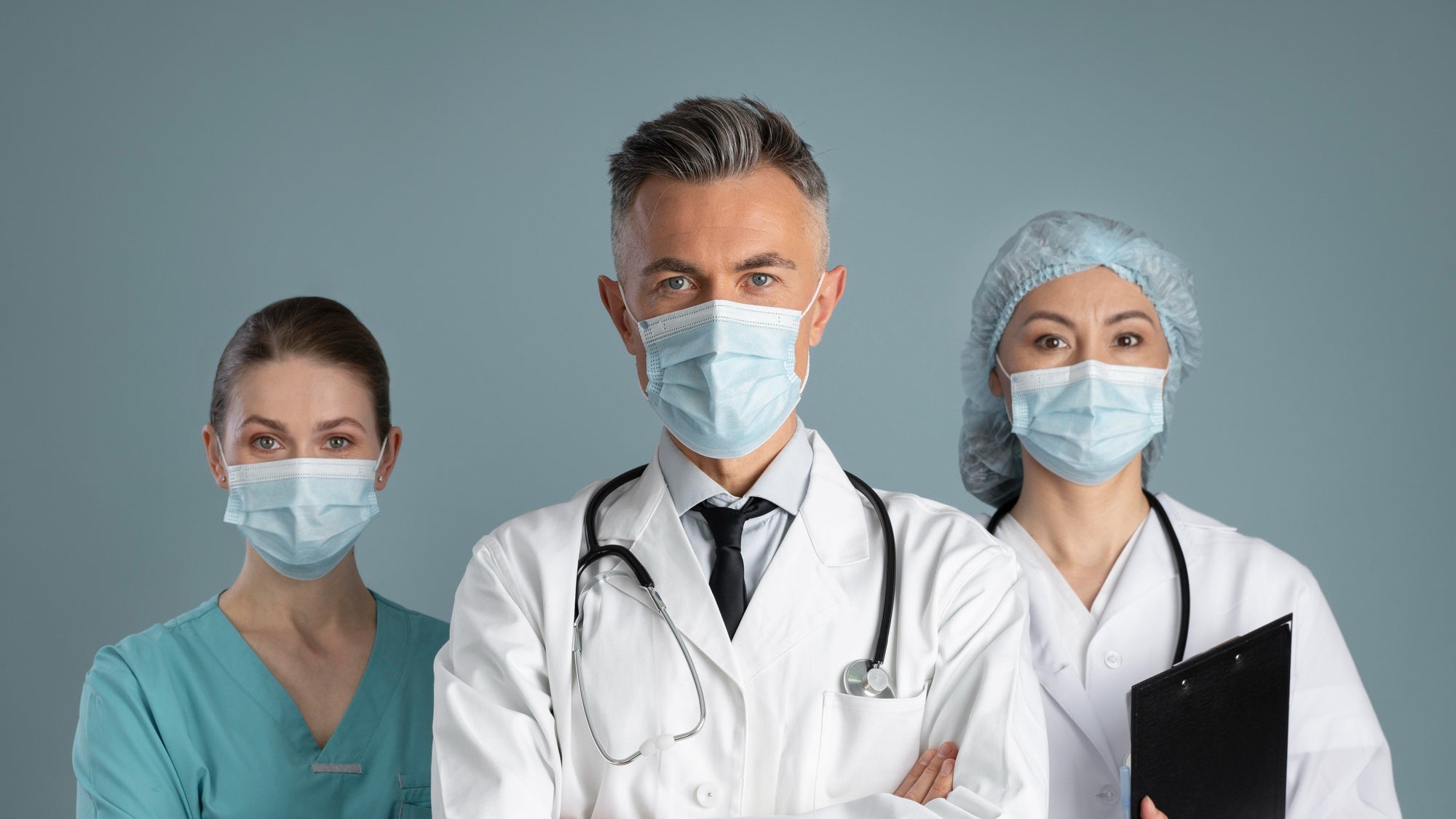 Three healthcare professionals wearing masks and uniforms against a gray background